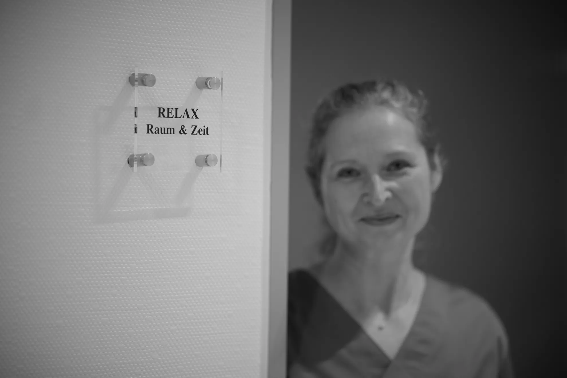 Raumschild "RELAX Raum & Zeit", im Hintergrund in etwas unscharf eine Person welche in die Kamera lächelt